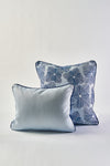 Two decorative pillows are shown against a plain background. In front is a light blue throw pillow; behind it is the Mod Flora Lumbar Pillow, showcasing blue floral and geometric patterns with dark piping along the edges.