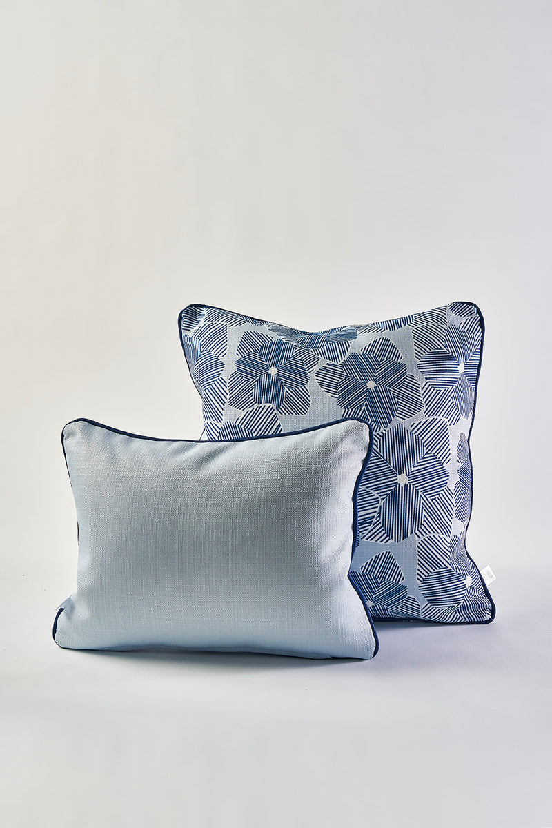 Two decorative pillows are shown against a plain background. In front is a light blue throw pillow; behind it is the Mod Flora Lumbar Pillow, showcasing blue floral and geometric patterns with dark piping along the edges.