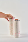 A hand holds a patterned Flamingo 20oz Tumbler with a straw next to a white stainless steel tumbler with pink flamingos, both featuring a no sweat exterior, on a white surface against a plain white background.