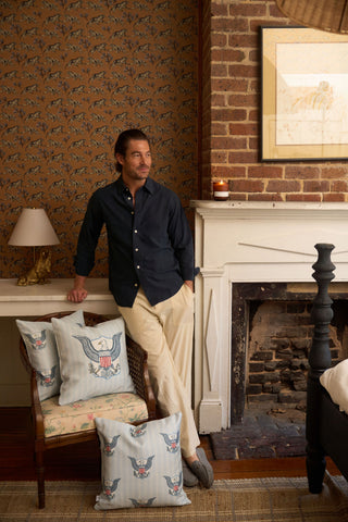 A man in a navy shirt and beige pants leans against a white fireplace mantel in a cozy room with patterned wallpaper, an exposed brick wall, and decorative One Nation Pillows featuring charming eagle designs.