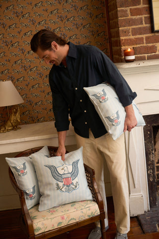 A man arranges a One Nation Pillow with a large eagle emblem in a cozy room featuring patterned wallpaper, a brick fireplace, a lit candle, and a vintage chair.