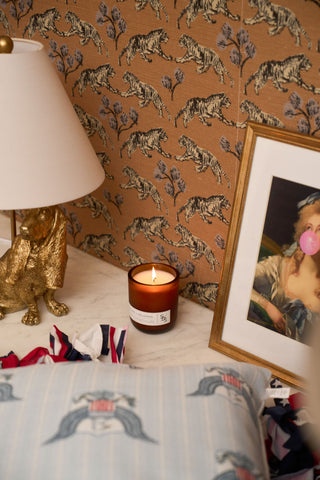 The Porches + Palomas Limited Edition Candle rests on a marble surface next to a gold animal lamp and a portrait of a woman blowing bubblegum, with tiger-patterned wallpaper and part of a decorative pillow visible.