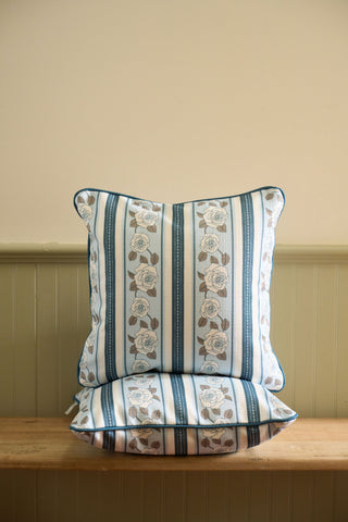 The Rose Garden Pillow is stacked with a striped pillow on a wooden bench against a light wall with wainscoting, bringing charm to the space.