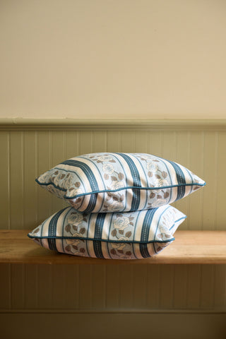 Two patterned pillows, including the Rose Garden Pillow in blue and beige, are stacked on a wooden bench set against a light-green paneled wall with a cream-colored upper section.