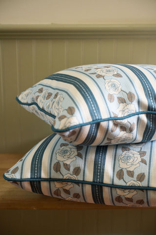 Two stacked cushions with blue and beige stripes and white rose designs, reminiscent of the Rose Garden Pillow, rest on a wooden surface against a light green paneled wall.