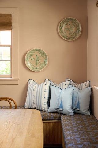 A cozy dining nook features a light wooden table, patterned bench, vintage floral pillow, and blue Set Sail Pillow. Sunlight streams in from the left window as three decorative plates hang on the beige wall above.