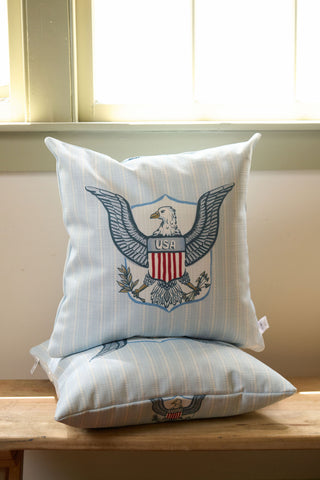 A pair of light blue striped One Nation Pillows, featuring an embroidered bald eagle with a USA shield, sits beside a vintage floral pillow from our Garden Prep Capsule Collection on a wooden bench by a sunlit window.
