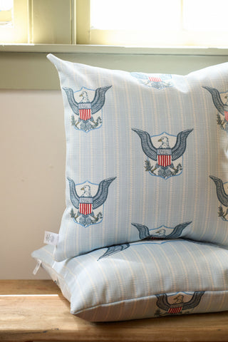 Two One Nation Pillows with light blue stripes, USA eagle emblems, and shields from the Garden Prep Capsule Collection are stacked on a wooden bench by a light green window.