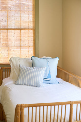A small bed with a white comforter and two light blue decorative pillows, including a Loyal Liberty Lumbar Pillow, sits by a window with a woven bamboo shade in a softly lit, neutral-colored room.