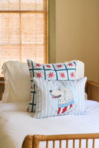 Two decorative pillows rest on a made bed: the Grandmillenial Garden Pillow with a blue and red star pattern, and the Loyal Liberty Lumbar Pillow featuring a dog in a patriotic scarf. Bamboo blinds cover the window in the background.