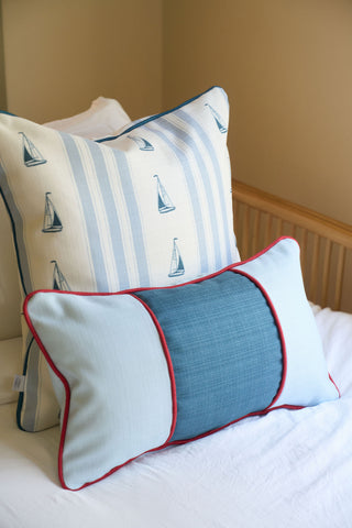 A white bed features two decorative pillows: the Stripes of the Union Extended Lumbar Pillow with blue and white stripes and sailboat prints at the back, and a light blue Grandmillenial Garden Pillow with darker blue center and red piping in front.