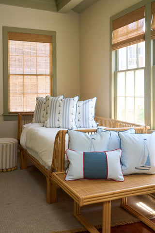 A cozy sunlit room with a rattan daybed and bench, decorated with the Stripes of the Union Extended Lumbar Pillow, vintage floral touches, neutral walls, woven window shades, and sailboat-patterned blue and white pillows.