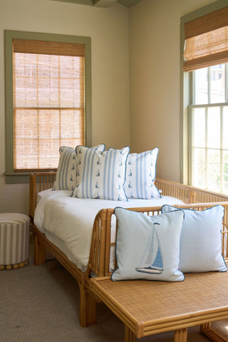 A cozy room features a rattan daybed with white bedding, the Set Sail Pillow in a blue sailboat motif, a vintage floral pillow, and sits by windows with woven shades. A striped round ottoman rests nearby on the light carpeted floor.