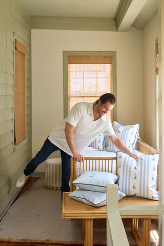 A man in a white polo and jeans arranges the Sea to Shining Sea Pillow among blue and white nautical pillows on a wooden bench in a bright, cozy room with pale green walls and bamboo-shaded windows.