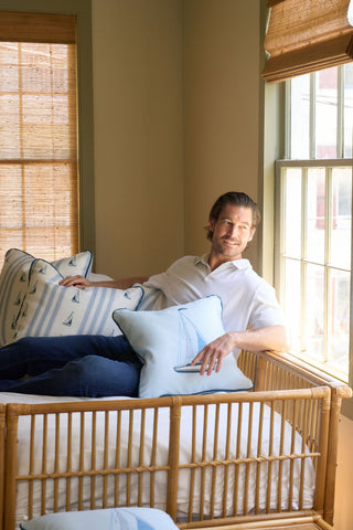A man with light brown hair and a beard sits on a rattan daybed by a sunny window, holding the Set Sail Pillow. The room’s light coastal décor includes a vintage floral pillow from the Garden Prep Capsule Collection.