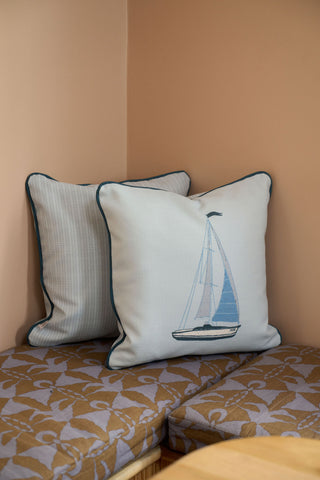 The Set Sail Pillow, featuring a sailboat design, rests on a bench beside a checkered pillow. A brown and lavender floral cushion and vintage floral pillow complete the charming seating arrangement.
