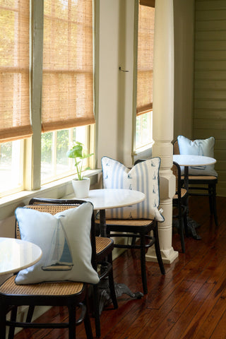 Small tables with white tops and wooden chairs line a sunlit room with large windows and bamboo shades. Blue and white sailboat-themed pillows, the Sea to Shining Sea Pillow, and a small plant decorate the space.