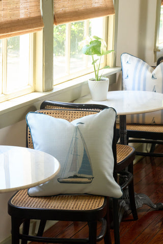 A cozy cafe corner with marble-topped tables, rattan chairs, a vintage floral pillow, the Set Sail Pillow, a striped pillow, a small potted plant, and large windows with bamboo blinds streaming in sunlight.
