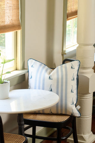 The Sea to Shining Sea Pillow, featuring blue sailboat and stripe designs, rests on a chair beside a round white table and window with bamboo blinds. A small potted plant on the table enhances this cozy, classic scene.