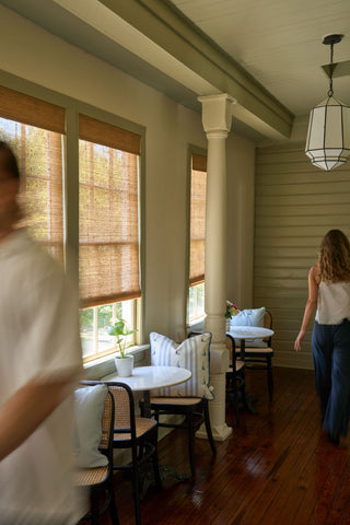 A cozy indoor seating area with small round tables, cushioned chairs, and bamboo window shades features the Sea to Shining Sea Pillow for added charm as warm light filters through windows onto wooden floors while two people move through the space.