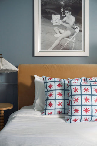 A bed with white sheets, two Star Spangled Pillows in a blue grid and red star pattern, a tan headboard, a round wooden side table, a pleated white lamp, and a black-and-white photo of an underwater person above the bed.