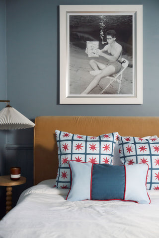 A cozy bedroom features a tan headboard, the Stripes of the Union Extended Lumbar Pillow with its bold patterns, two additional pillows, a bedside table with a candle, and a quirky black-and-white photo of someone reading underwater above the bed.