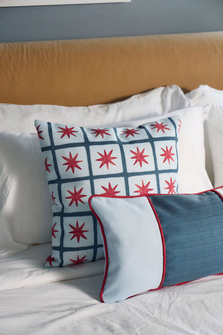 A bed with white sheets and pillows features the Star Spangled Pillow, showcasing a blue grid and red star pattern, next to a Grandmillennial Garden Pillow. A tan headboard is in the background.