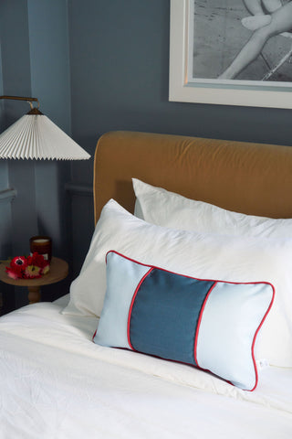A neatly made bed with white sheets and a Stripes of the Union Extended Lumbar Pillow. A side table displays a lamp and tray of red flowers, while a framed photo hangs on the dark blue wall above the bed.