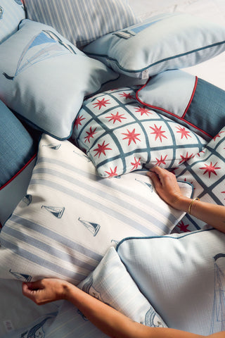 A person arranges a pile of decorative pillows, featuring the Sea to Shining Sea Pillow with sailboats, stripes, and starburst designs in blue, white, and red, paired with vintage floral and geometric patterns.