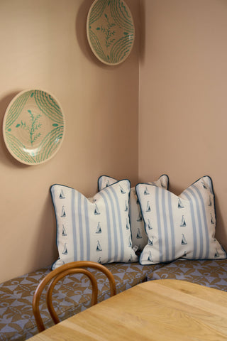 A cozy corner featuring beige walls, a wooden chair, a light wood table, striped sailboat pillows on a cushioned bench, and the Sea to Shining Sea Pillow from the Garden Prep Capsule Collection adds charm beside decorative plates.