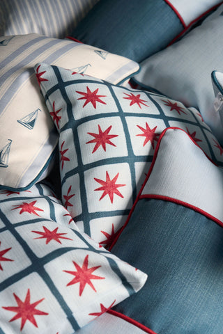A close-up of decorative pillows, featuring a Grandmillenial Garden Pillow alongside nautical-themed designs such as the Star Spangled Pillow with red stars, plus blue stripes, sailboats, and blue trim arranged in a cozy, overlapping display.