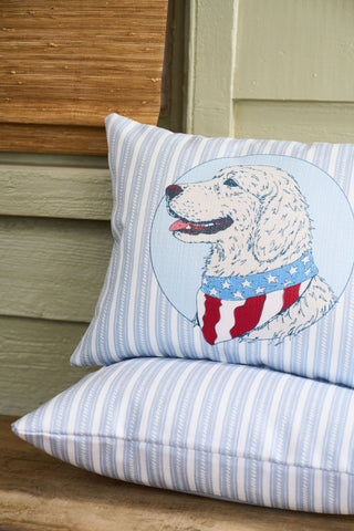 The Loyal Liberty Lumbar Pillow, with its blue-and-white stripes and dog illustration in a stars-and-stripes bandana, adds charming garden-inspired decor to a wooden bench alongside a vintage floral pillow.