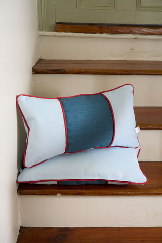 The Stripes of the Union Extended Lumbar Pillows from the Garden Prep Capsule Collection, with light blue fabric, a dark blue center panel, and red piping, are stacked on wooden stairs by a white wall.