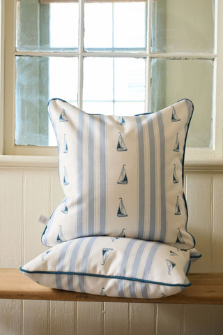 Two Sea to Shining Sea Pillows, with blue and white stripes and sailboat patterns, are stacked on a wooden bench by a window with divided panes.