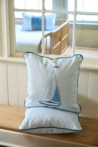 Two light blue pillows with navy trim, including the Set Sail Pillow embroidered with a sailboat, sit on a wooden bench. Through the window, a bed with blue pillows and a Grandmillenial Garden Pillow is visible in the background.