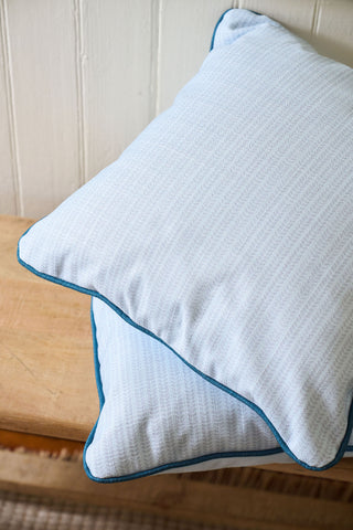 Two Set Sail Pillows from the Garden Prep Capsule Collection, featuring white fabric with blue edging, are stacked on a wooden bench against a white paneled wall.