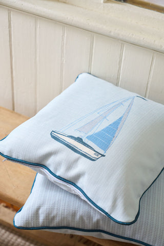 The Set Sail Pillow, featuring a blue sailboat design, is stacked on a light blue cushion atop a wooden bench with a white paneled wall in the background.