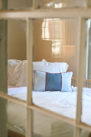 A cozy bed with white pillows and a Stripes of the Union Extended Lumbar Pillow is seen through a window with white panes. Soft natural light filters in, adding vintage charm and creating a calm, inviting atmosphere.
