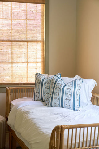 A cozy bedroom features a bamboo daybed with white bedding, a Rose Garden Pillow, and a blue and white striped cushion. Woven blinds on the window let in natural light, enhancing the room’s neutral tones.