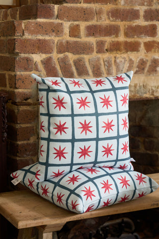 Two square Star Spangled Pillows with a blue grid and red starburst pattern are stacked on a wooden bench in front of a brick wall and fireplace.