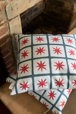 Two white Star Spangled Pillows, featuring a grid of dark blue lines and red eight-pointed stars, rest on a wooden surface in front of a brick fireplace.