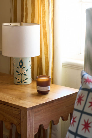 A wooden side table with a floral lamp, a Porches + Palomas Limited Edition Candle in a brown glass holder, and a patterned pillow sits by a window with yellow and white striped curtains inspired by Charleston, SC.