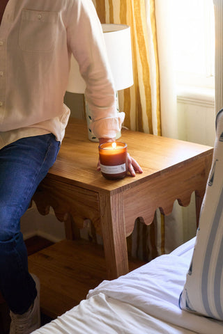 A person in a light shirt and jeans sits on a bed, resting their hand on a wooden nightstand next to the Porches + Palomas Limited Edition Candle. A lamp and striped curtains are visible by the sunlit window.