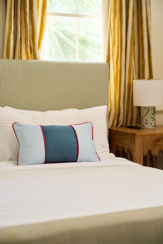 A neatly made bed with white linens, a Stripes of the Union Extended Lumbar Pillow as an accent, a striped headboard, yellow striped curtains, and a wooden nightstand with a lamp by the window.