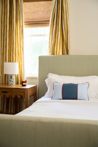 A neatly made bed with white bedding features the Stripes of the Union Extended Lumbar Pillow. A wooden nightstand with a lamp and candle sits beside it, while yellow striped curtains frame the window behind the bed.