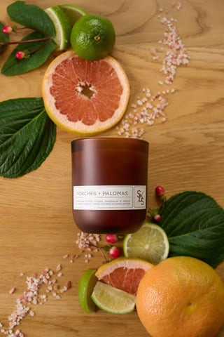 The Porches + Palomas Limited Edition Candle, a hand-poured brown soy candle from Charleston, rests on wood among grapefruit, whole and sliced limes, green leaves, pink berries, and pink salt—evoking a refreshing citrus lychee aroma.