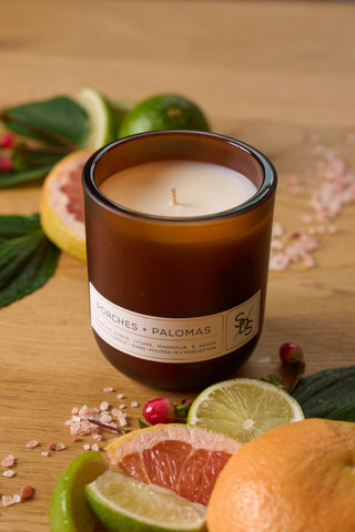 A clean-burning Porches + Palomas Limited Edition Candle sits on a wooden surface, surrounded by grapefruit and lime slices, green leaves, pink salt, and small red berries.