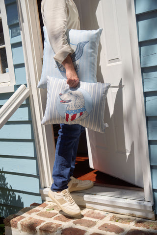 A person in jeans and cream sneakers carries two decorative pillows, including a Grandmillenial Garden Pillow with animal illustrations and the Loyal Liberty Lumbar Pillow, entering a light blue house through a white door.