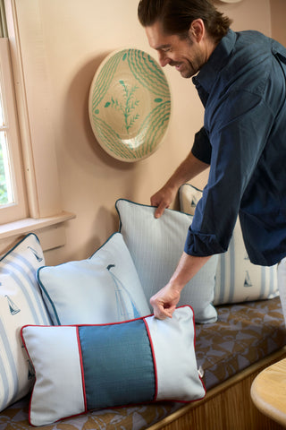 A man arranges blue and white throw pillows, including the Stripes of the Union Extended Lumbar Pillow, on a cushioned bench beneath a window. A decorative bowl hangs on the wall above the bench.