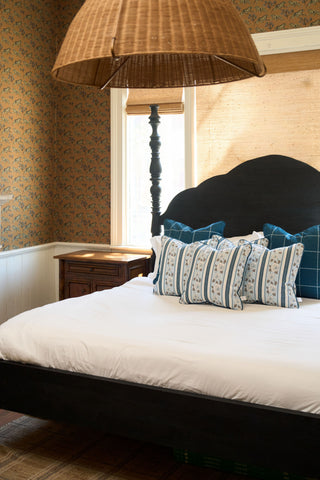 A cozy bedroom with a black wooden bed, white bedding, blue and white striped pillows, the Rose Garden Pillow, wooden nightstand, wicker pendant light, floral wallpaper, and sunlight streaming through a window with a woven shade.
